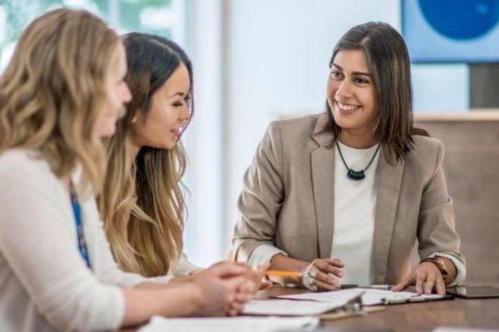 Três mulheres em um ambiente de trabalho, com uma delas sorrindo e olhando para a colega, enquanto as outras duas prestam atenção. A imagem ilustra a prática de escuta ativa durante uma reunião.