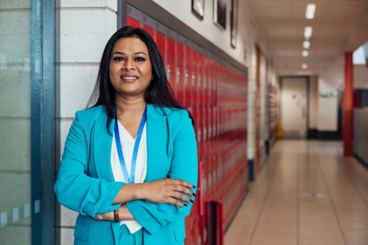 Uma mulher sorridente, posa com os braços cruzados no corredor de uma escola, com armários vermelhos ao fundo. Ela representa a liderança na gestão escolar, transmitindo confiança e profissionalismo.