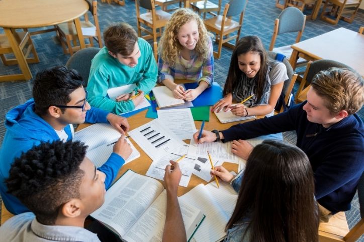 Um grupo de estudantes sentados ao redor de uma mesa em uma sala de aula, interagindo e discutindo enquanto apontam para um livro aberto. Eles parecem estar envolvidos em uma atividade colaborativa.