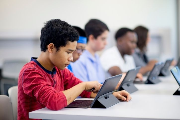 Jovens concentrados utilizando laptops em uma sala de aula, em uma mesa compartilhada.