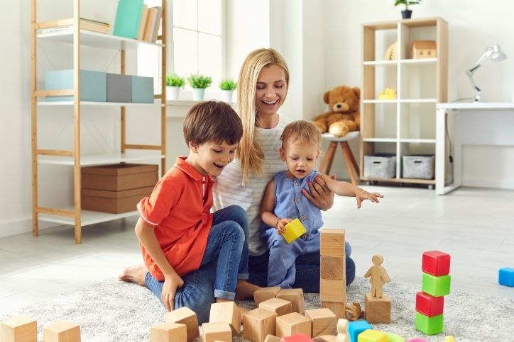 Mãe brincando com dois filhos pequenos em um ambiente doméstico, construindo torres com blocos de madeira e coloridos. Cena de aprendizado, diversão e conexão familiar.