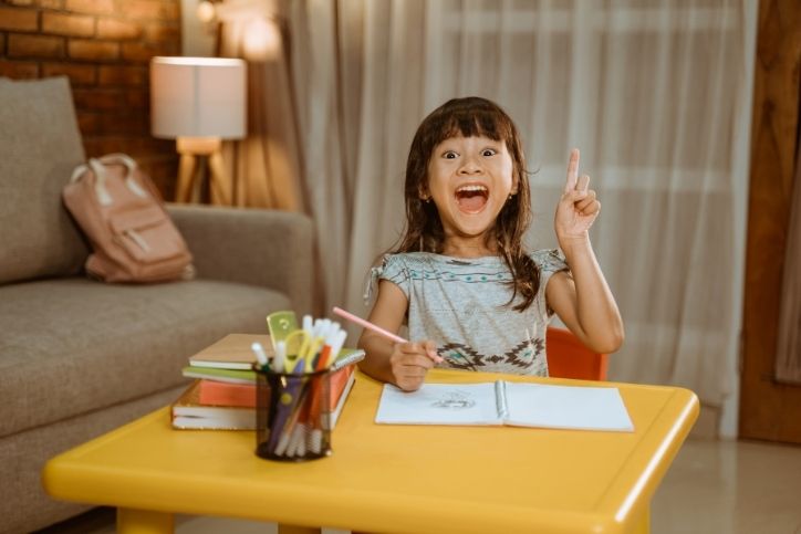 Uma menina sorridente, segurando um lápis, empolgada ao lado de seus materiais escolares em uma mesa amarela, com uma mochila e sofá ao fundo.