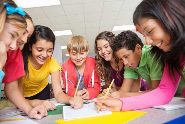 Um grupo de estudantes em uma sala de aula, sorrindo e trabalhando juntos em um projeto, com papéis e lápis sobre a mesa.