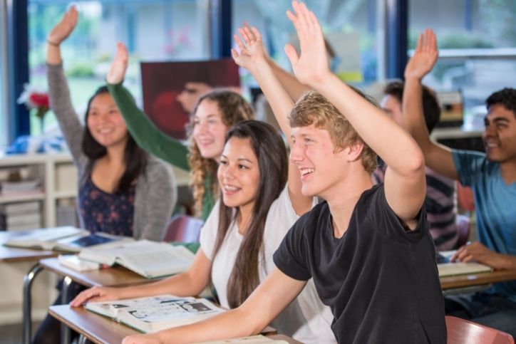 Um grupo de adolescentes levanta as mãos em uma sala de aula, interagindo de forma animada com a aula.