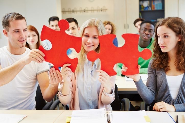 Estudantes em sala de aula colaborando ao montar peças de um quebra-cabeça vermelho, representando trabalho criativo em equipe.