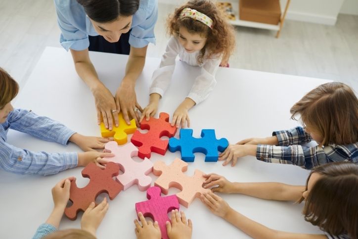 grupo de crianças e professora montam um quebra-cabeça em equipe.