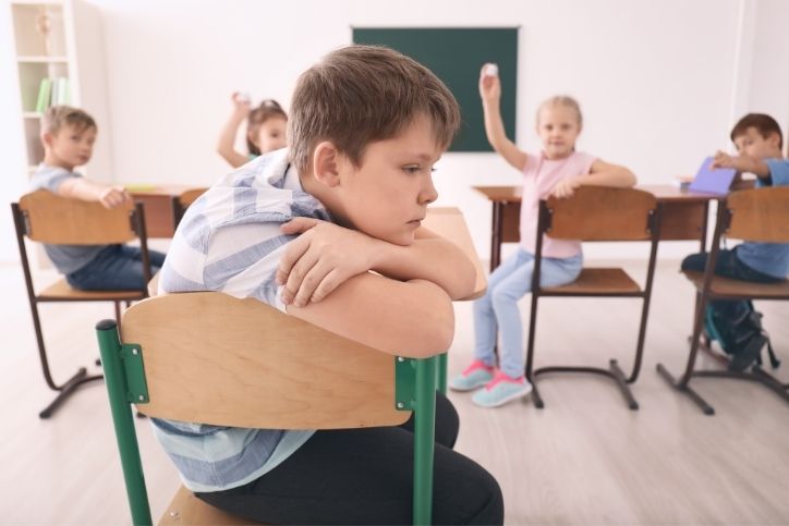 criança isolada na sala de aula sofrendo bullying dos colegas ao fundo.