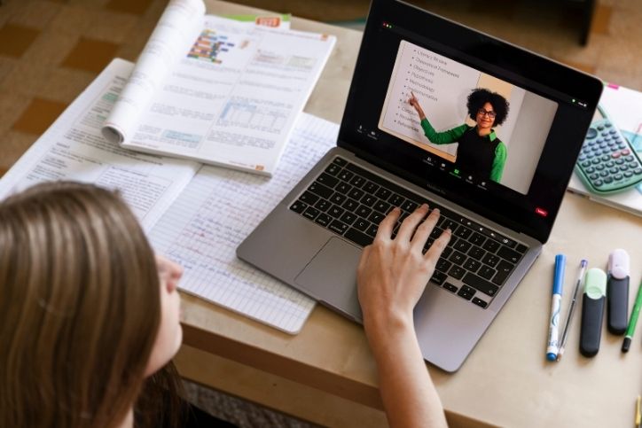 aluna assiste a aula online no notebook com caderno e livros abertos.