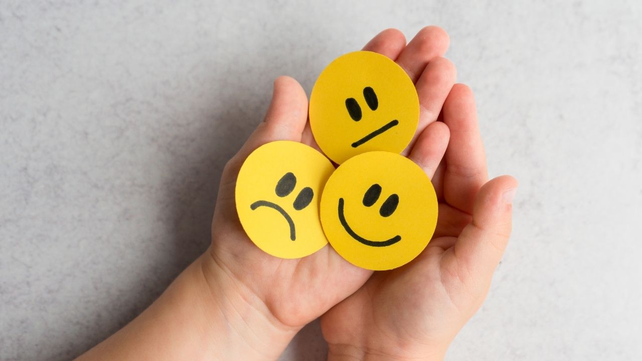 mãos segurando emojis de carinhas felizes, tristes e neutras representando emoções.