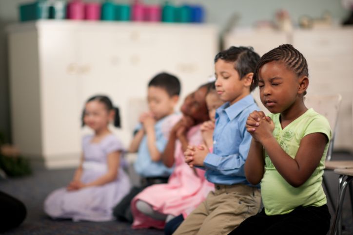 crianças sentadas com olhos fechados e mãos postas em gesto de oração.