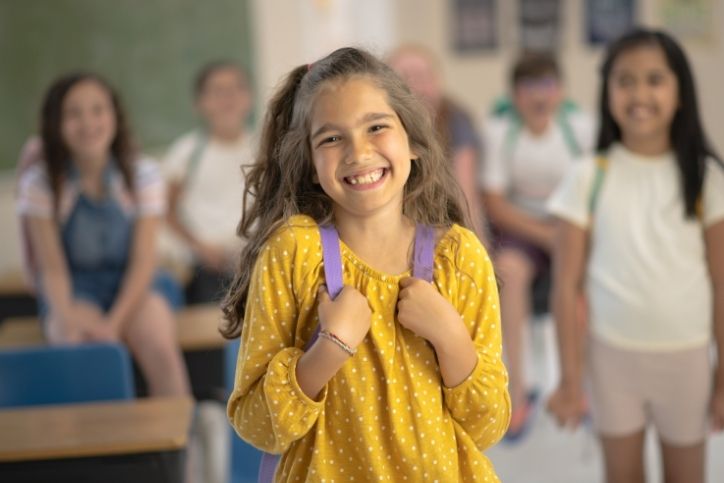criança sorrindo com mochila nas costas dentro da sala de aula.