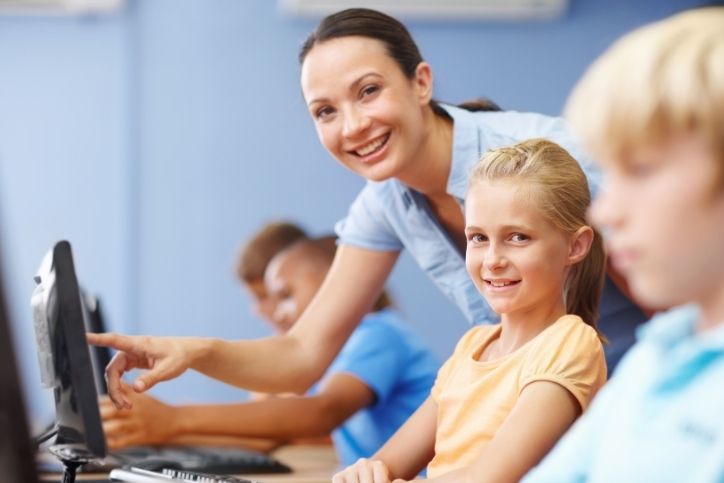 Professora auxiliando aluna em sala de informática escolar, com crianças utilizando computadores como recursos didáticos.