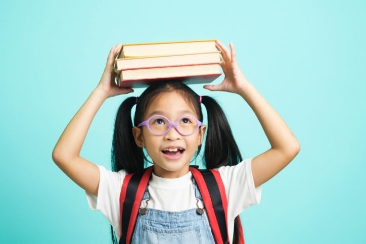 menina sorridente com óculos roxos e mochila segura três livros empilhados na cabeça com fundo azul claro.